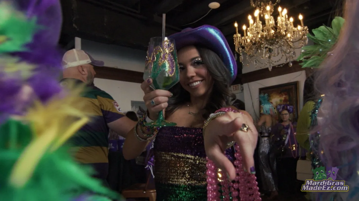 A guest in a sequined dress and purple hat raising a decorated goblet at a Mardi Gras ball