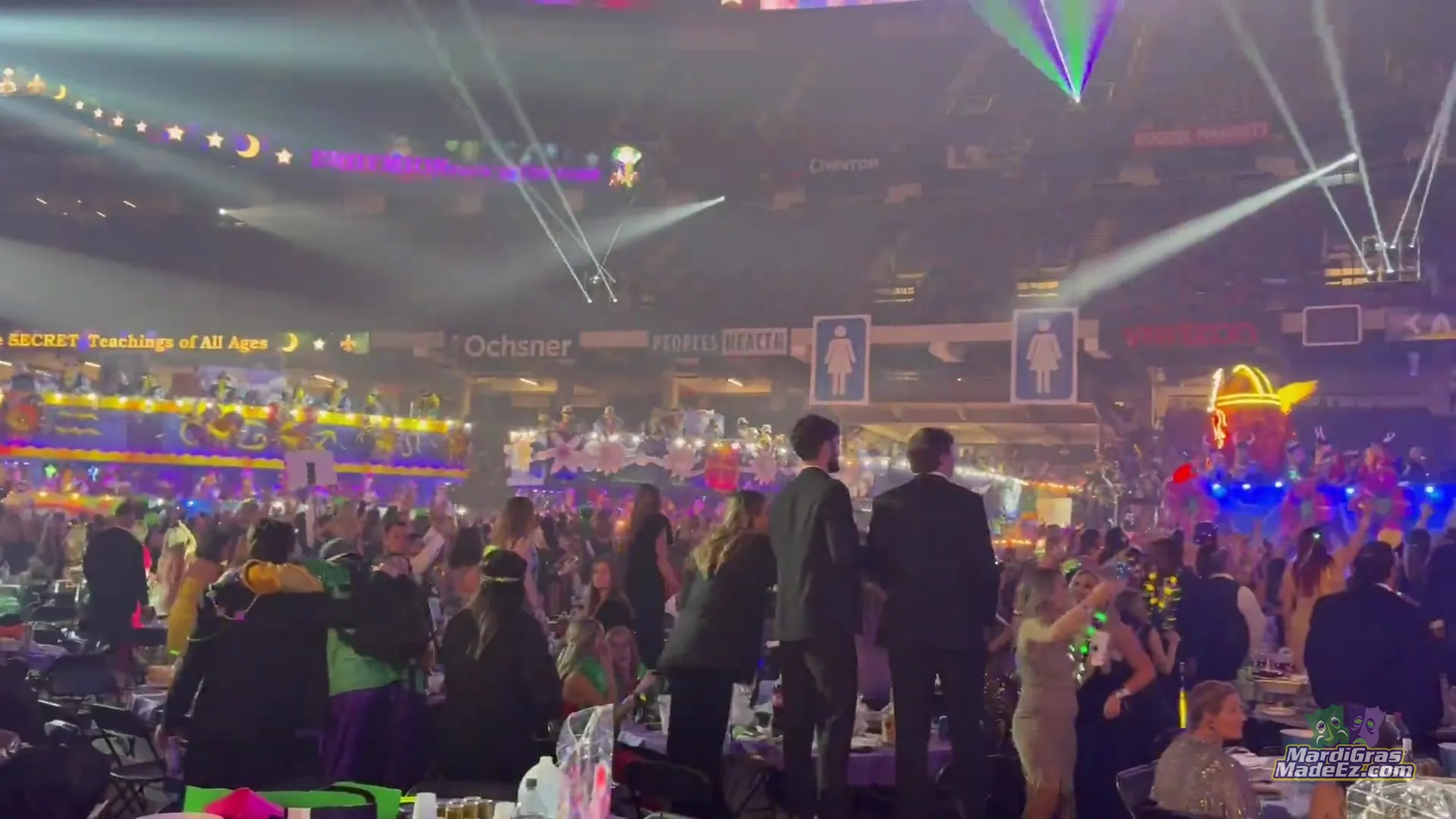 Guests in evening wear at their tables inside an arena as lit floats pass under laser lights